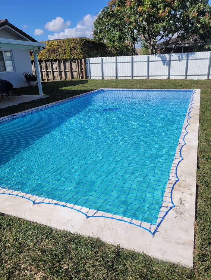 Una piscina cristalina con red de seguridad sobre un césped verde y una cerca blanca detrás